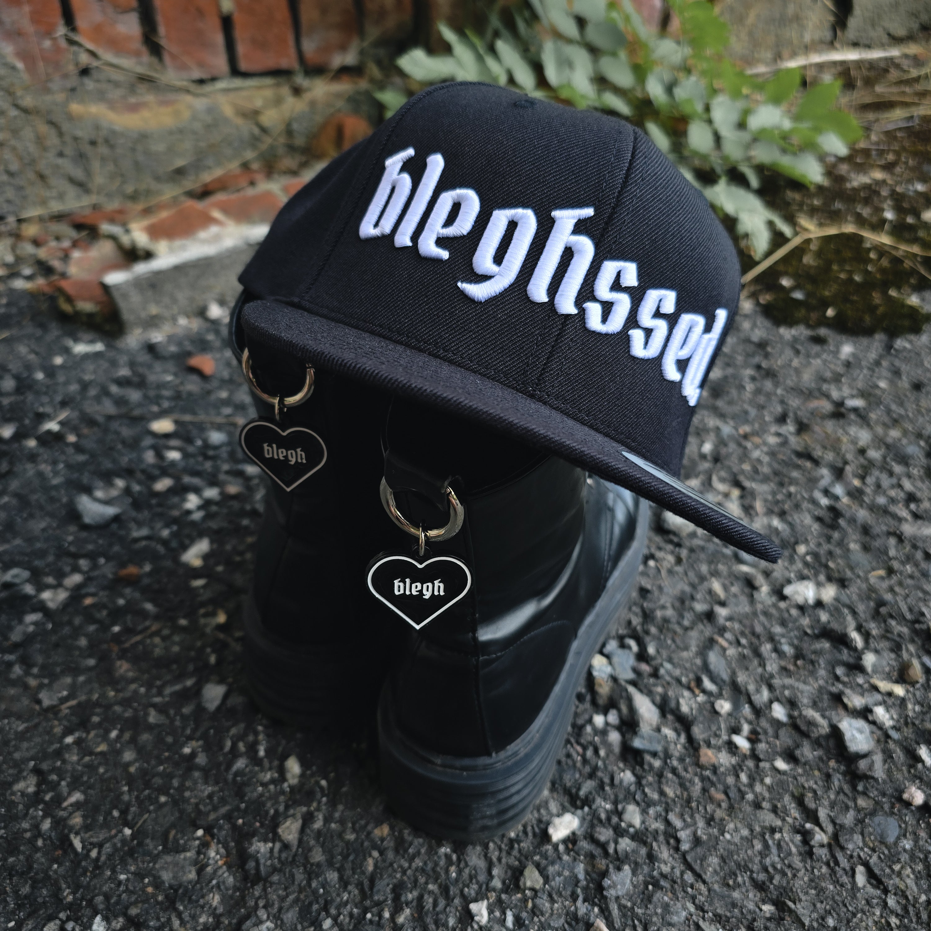 Snapback Cap mit bleghssed-Stickerei, hängend auf schwarzen Stiefeln mit blegh Boot Charms – Streetwear Accessoires für die Metalcore-Szene