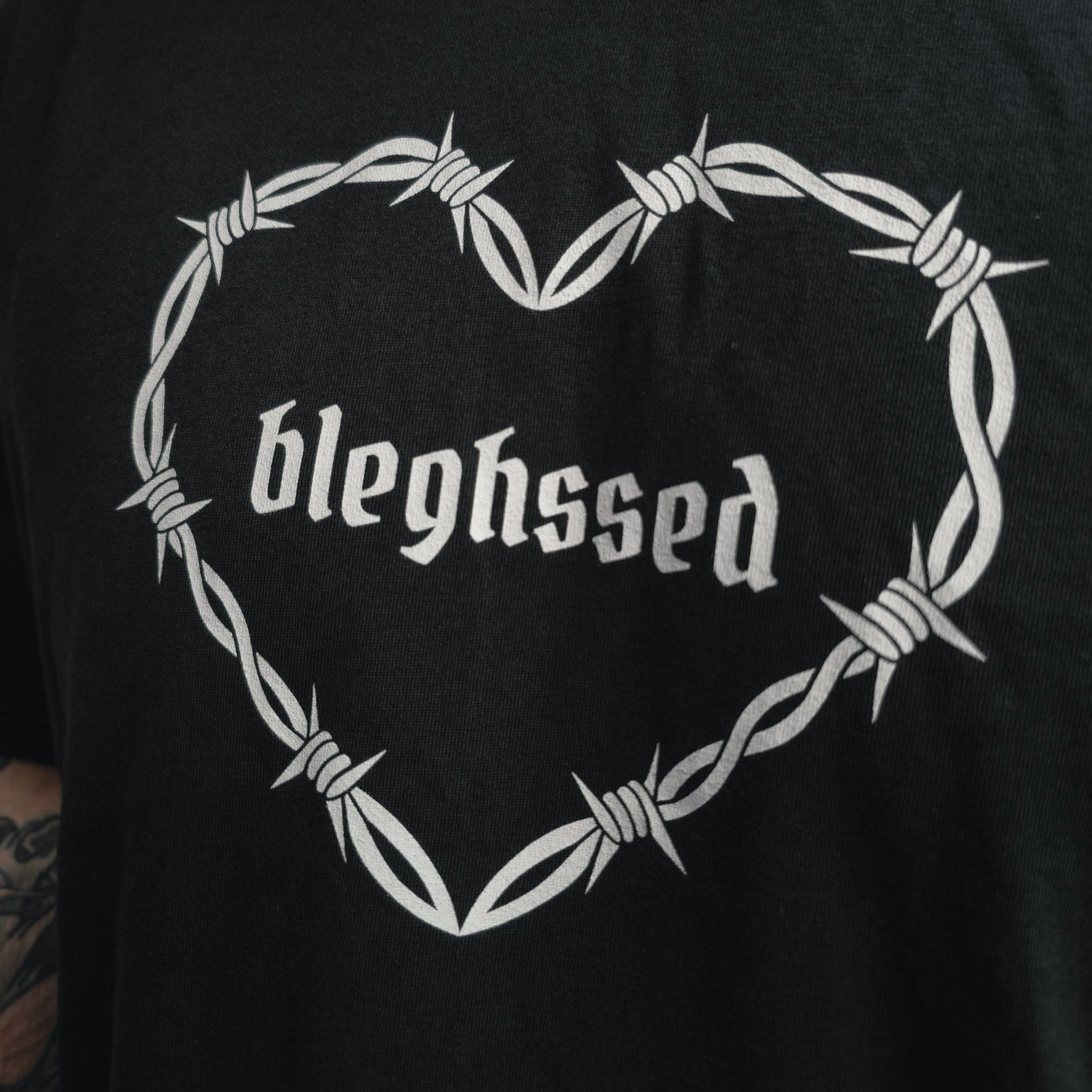 Nahaufnahme des „bleghssed“-Schriftzugs im Herz aus Stacheldraht auf schwarzem T-Shirt